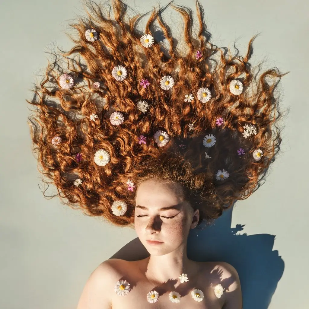Woman with ginger wavy hair, eyes shut, lying on the floor with flowers spread in her hair